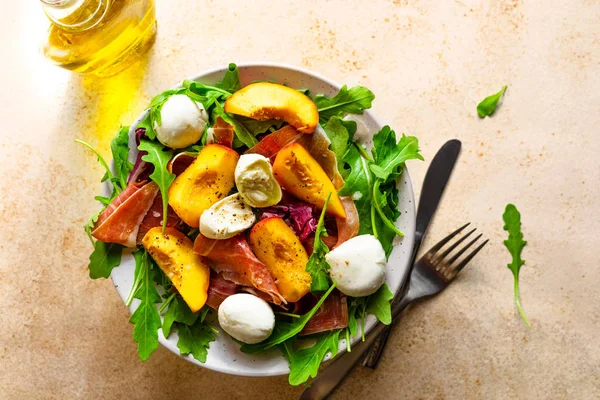 Light salad of fresh peaches, parma ham, mozzarella and green salad mix, olive oil, fork and knife.