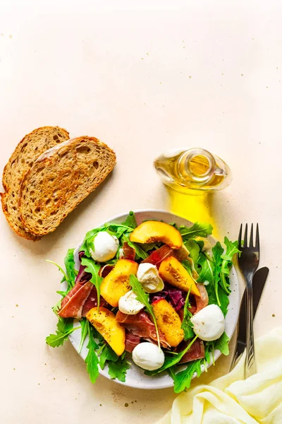 Light salad of fresh peaches, parma ham, mozzarella and green salad mix, olive oil, bread, fork and knife.