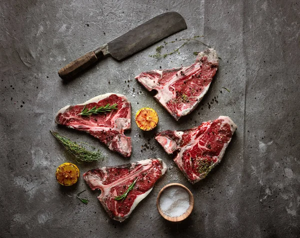 Filetes de hueso en t crudos, cuchillo de carnicero, condimento y mitades de limón a la parrilla sobre fondo texturizado de hormigón gris, vista superior — Foto de stock gratis