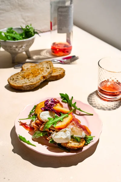 Parma skinka, mozzarella och persikor smörgås på en tallrik, skål med arugula, lite bröd och Rosen vin sköt med hårt ljus — Stockfoto