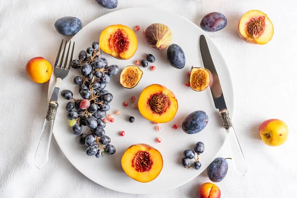 Setengah buah persik, nektarin, anggur, ara di piring putih, sendok makan di taplak meja putih . — Stok Foto
