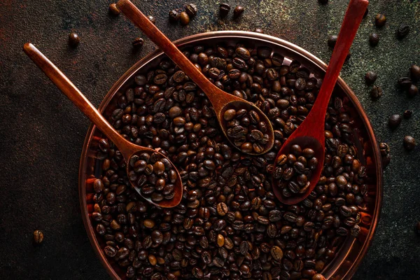 Round copper tray, wooden spoons full of kopi luwak coffee beans on dark background, top view
