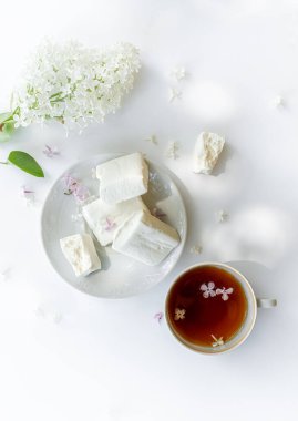 Ev yapımı marşmelov, çay, beyaz arka planda beyaz leylak dalı. Işıklar ve gölgelerle çekilmiş açık hava çekimi. Üst görünüm, portre yönelimi