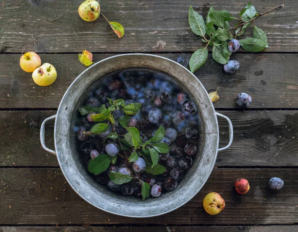 Plums soaking in water in a small tub, apples on old wooden background — Stock Photo, Image