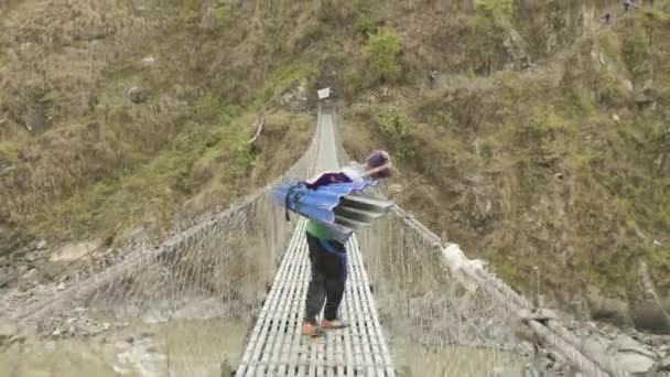МАНАСЛУ, НЕПАЛ - Март 2018 года: Портер с тяжелыми рюкзаками проходит через подвешенный мост . — стоковое видео