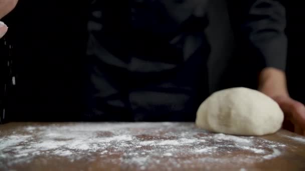 Повар посыпает муку на деревянный стол на черном фоне — стоковое видео