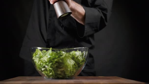 Повар посыпает соль и специи в зеленый салат на черном фоне — стоковое видео