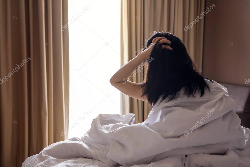 a Lazy Woman with Messy Hair sitting on White Bed and Looking Outside Through Window in the Morning