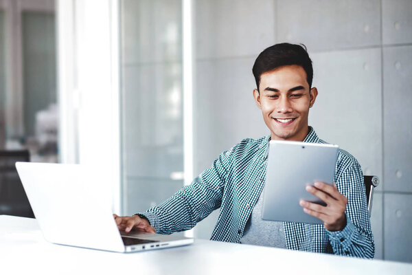 Happy Young Asian Businessman Working on Computer Laptop in his Workplace. Smiling and looking at Digital Tablet