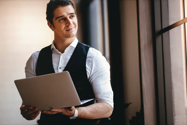 一位年轻的白种人商人在现代工作场所的电脑笔记本电脑上工作 微笑的商人站在窗边向外面看 — 图库照片