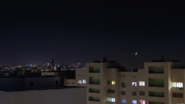 Gece şehir timelapse 4k. Yıldızlar ve yeni ay gökyüzünde hareket ediyor. Konut daireleri pencereleri yanar ve kapanır. Yüksek katlı binalarla şehir silueti
