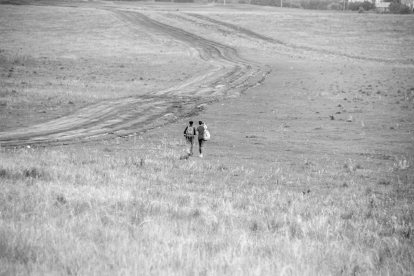 Dvě mladé dívky s batohy a fotolaboratoře dělají na telefonu na venkovské cestě. Pastvina pro koňské farmy s klisny a foalem. Malá vesnička se starými domy. Letní krajina se zelenými kopci — Stock fotografie