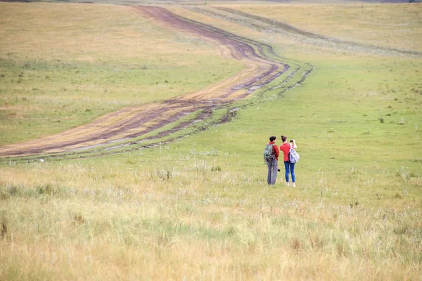 Duas meninas com mochilas e câmera fotográfica fazem selfie no telefone perto da estrada rural. Horse farm pasto com égua e potro. Paisagem de verão com colinas verdes — Fotografia de Stock