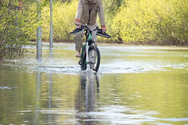 Велосипедист с голыми ногами пытается преодолеть воду во время наводнения весной. Дорога под водой, как река лопнула его берега — стоковое фото