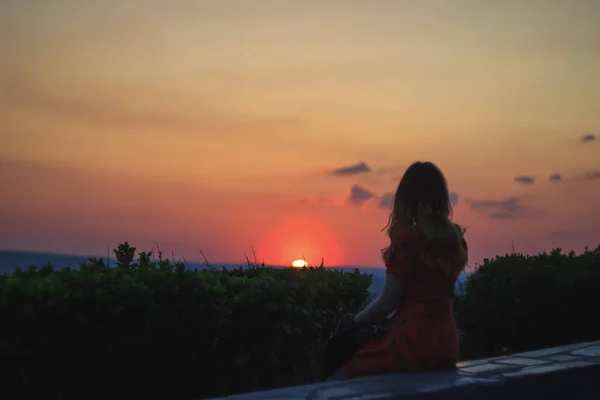 Silueta de una mujer vestida sentada sobre piedra en la cima de la colina. Ella ve el círculo rojo del sol cayendo — Foto de Stock