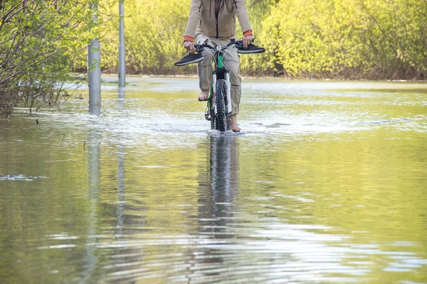 Велосипедист с голыми ногами пытается преодолеть во время наводнения весной. Дорога под водой, как река лопнула его берега — стоковое фото
