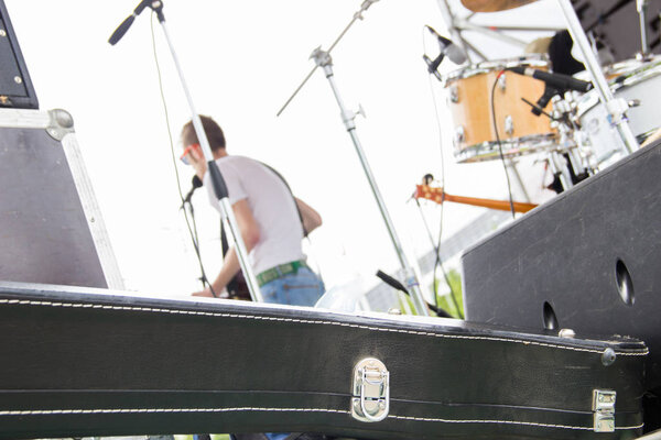 Музыкальная группа играет на открытом воздухе на летнем фестивале, вид из-за сцены, черный чехол для гитары, вокалист на сцене
