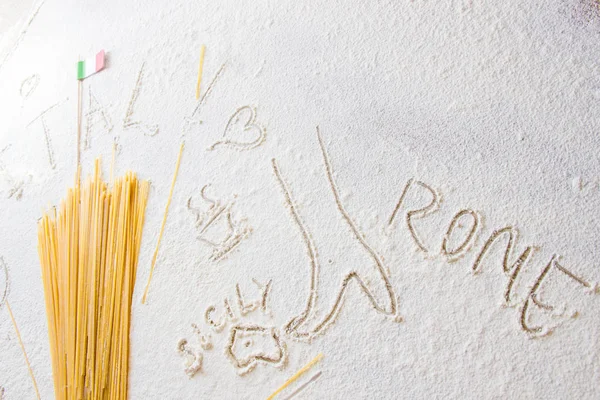 Uncooked pasta spaghetti macaroni and italian flag on floured white background. Words Italy, Rome, Sicily written on flour from hand, hand drawn Italy map silhouette, a cup of coffee, heart