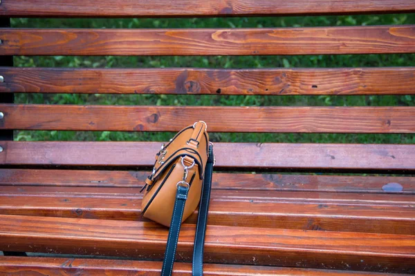 A close-up of a leather fashionable lady\'s bag, made in brown color. She is standing on an old bench in a summer park