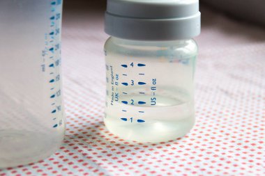 Masanın üzerinde pembe desenli masa örtüsüyle kaplı bebek aksesuarları. Karanlık arka planda suyla birlikte ölçekli besleme şişeleri. Küçük ve büyük şişelerin fotoğrafını kapat.