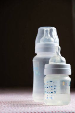 Masanın üzerinde pembe desenli masa örtüsüyle kaplı bebek aksesuarları. Karanlık arka planda ölçekli besleme şişeleri. Küçük ve büyük şişelerin fotoğrafını kapat.