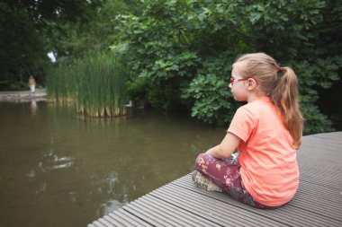 Genç kız köprüde oturur ve gölete bakar.