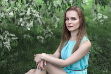 Mavi elbiseli genç sarışın bir kızın portresi. O çimlerin üzerinde oturuyor. Çiçekli ağaçlar.