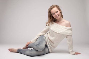 Güzel genç kız beyaz arka planda poz veriyor ve oturuyor. Beyaz kazaklı sarışın..