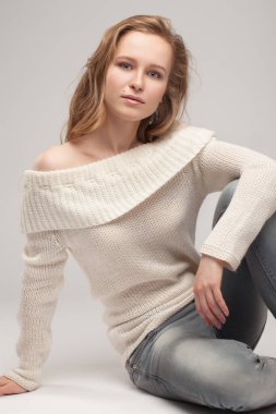 Güzel genç kız beyaz arka planda poz veriyor ve oturuyor. Beyaz kazaklı sarışın..