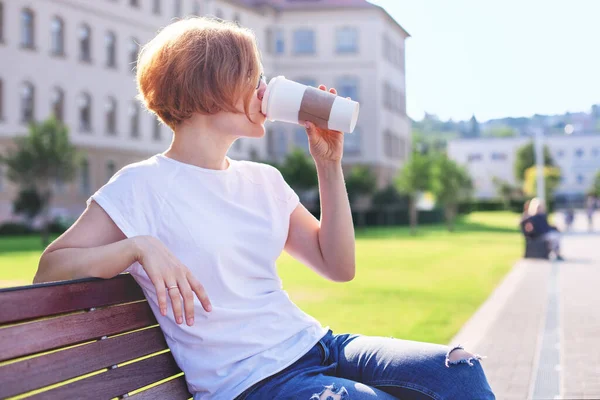 Beyaz tişörtlü ve kot pantolonlu bir kız bankta oturur ve beyaz bir bardaktan su içer. dinlenme ve sessizlik. Hayatın tadı.