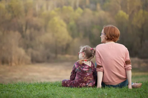 Mom and daughter in glasses are sitting on the grass and watching the distance against the backdrop of a beautiful forest