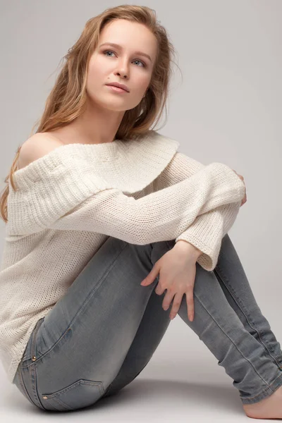 Güzel genç kız beyaz arka planda poz veriyor ve oturuyor. Beyaz kazaklı sarışın..