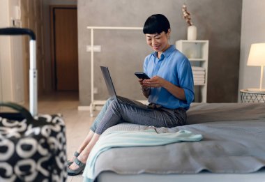 Yatakta dizüstü bilgisayar ve akıllı telefon kullanan bir kadın gülümsüyor.