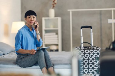 Yatakta oturan bir kadın yanında bavuluyla telefonla konuşuyor.