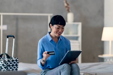 Smiling woman sitting on bed holding credit card using tablet computer