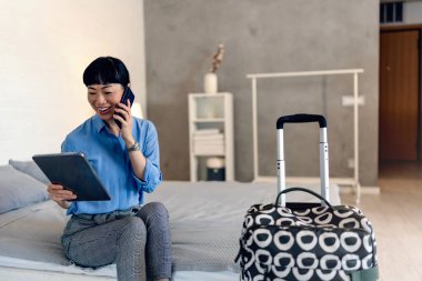 Woman sitting on bed using tablet while talking phone near suitcase