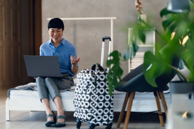 Smiling woman sitting on bed using laptop near patterned suitcase