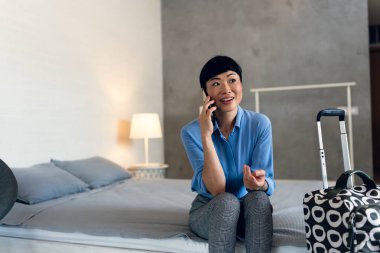 Smiling woman sitting on bed talking using smartphone indoors