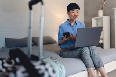 Smiling woman sitting on bed using laptop and smartphone