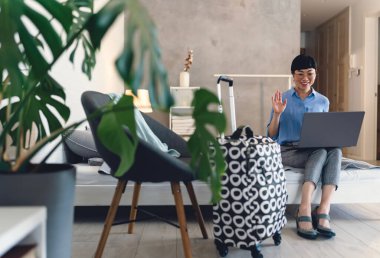 Smiling woman sitting on bed using laptop waving near suitcase indoors