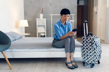 Smiling woman sitting on bed checking phone near suitcase