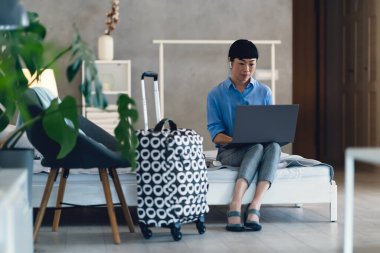 Woman sitting on bed using laptop near patterned suitcase indoors