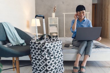 Smiling woman sitting on bed using laptop while packing suitcase nearby