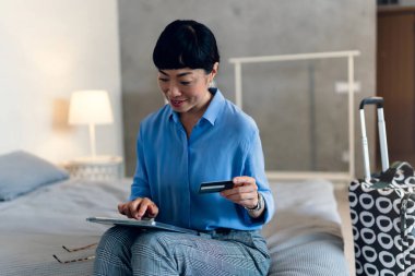 Smiling woman sitting on bed holding credit card using laptop computer shopping