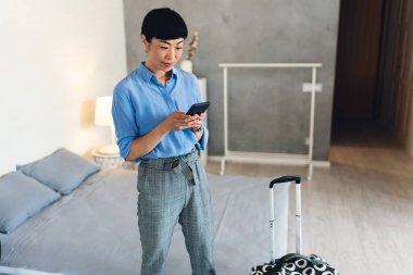 Young woman standing near bed using smartphone beside suitcase indoors