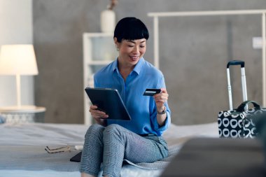 Smiling woman sitting on bed holding tablet and credit card