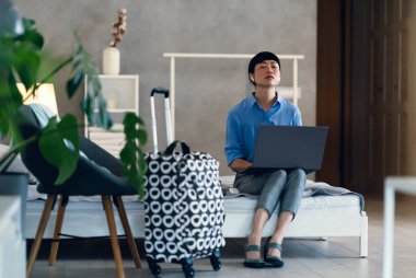 Woman sitting on bed using laptop near suitcase relaxing indoors