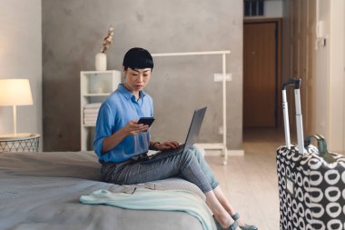 Woman sitting on bed using laptop and smartphone indoors