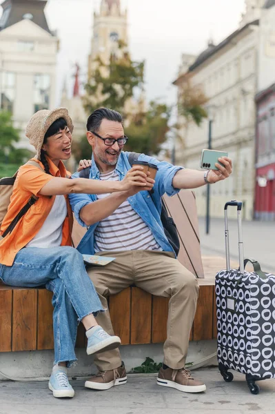 Smiling couple sitting outdoors taking cheerful selfie together with coffee cups