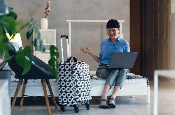 Smiling woman sitting on bed using laptop near suitcase indoors